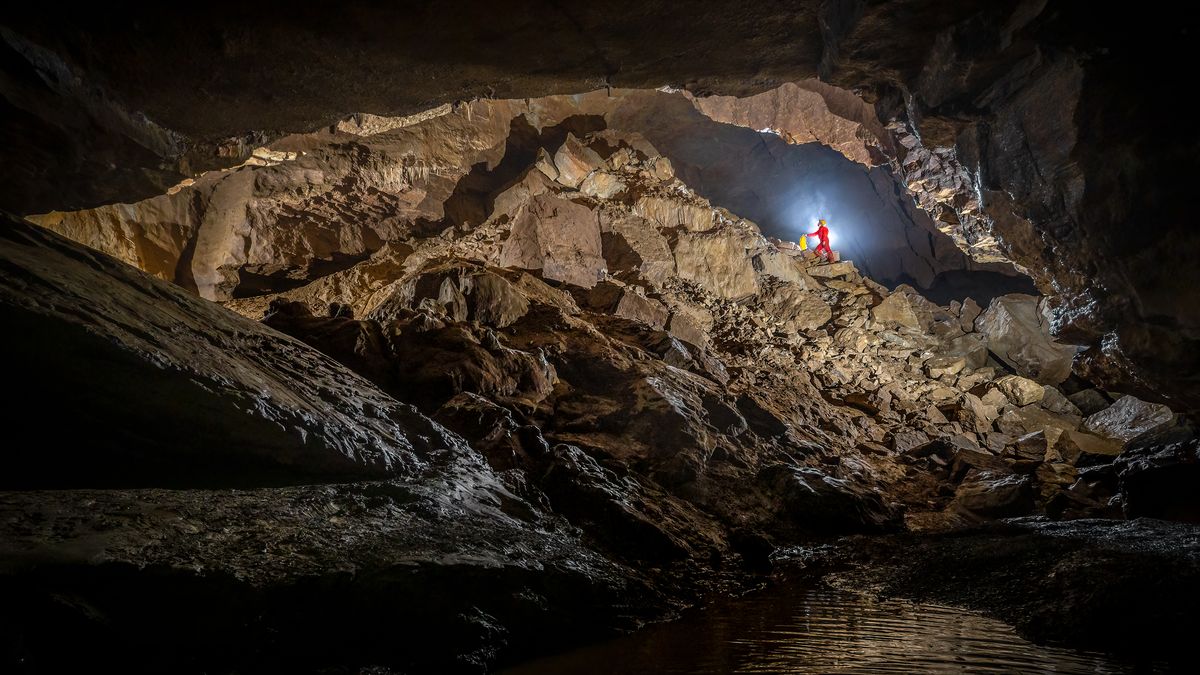Spéléologue en combinaison rouge escaladant un vaste éboulis de blocs, au fond d'une grande salle souterraine.